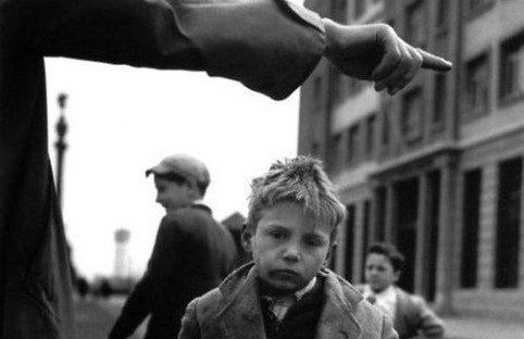 Un enfant triste se tient au premier plan, observant un homme qui pointe du doigt.