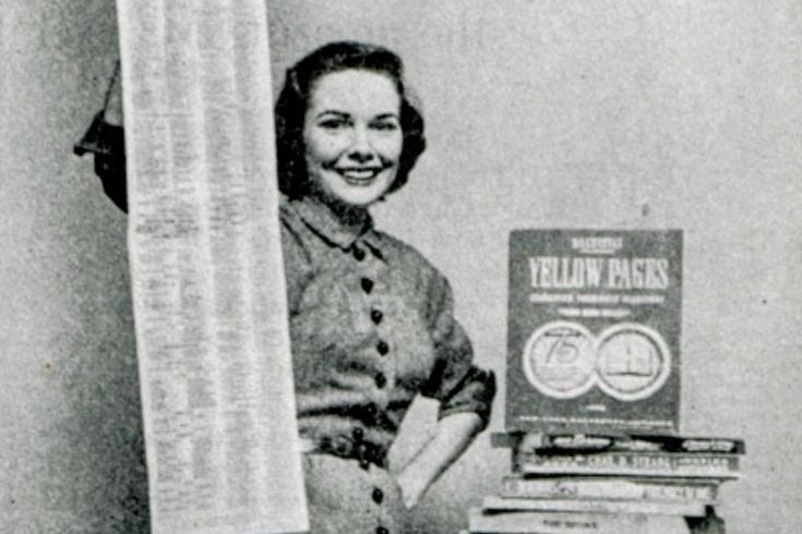 Femme souriante, tenant un rouleau de papier devant une pile de livres et un manuel.